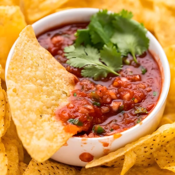 A tortilla chip dipped into a white bowl of fresh salsa, surrounded by more chips.